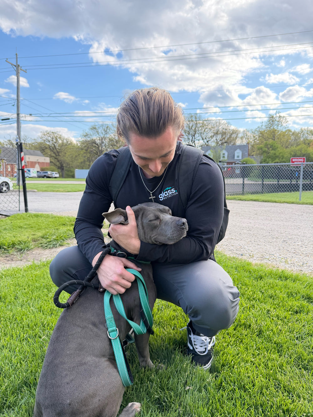Man kneeling on grass with his head bowed toward a gray pit bull on a teal leash.