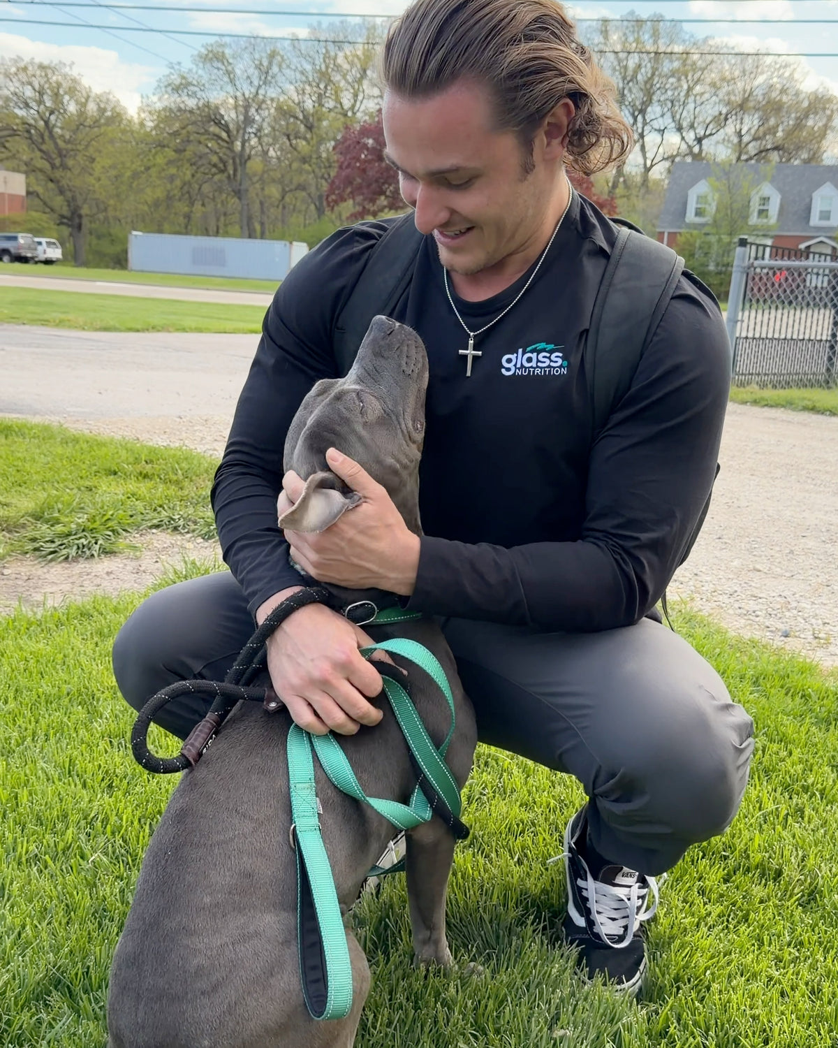 Gray pit bull on a teal leash nuzzling the face of a man kneeling on the grass.