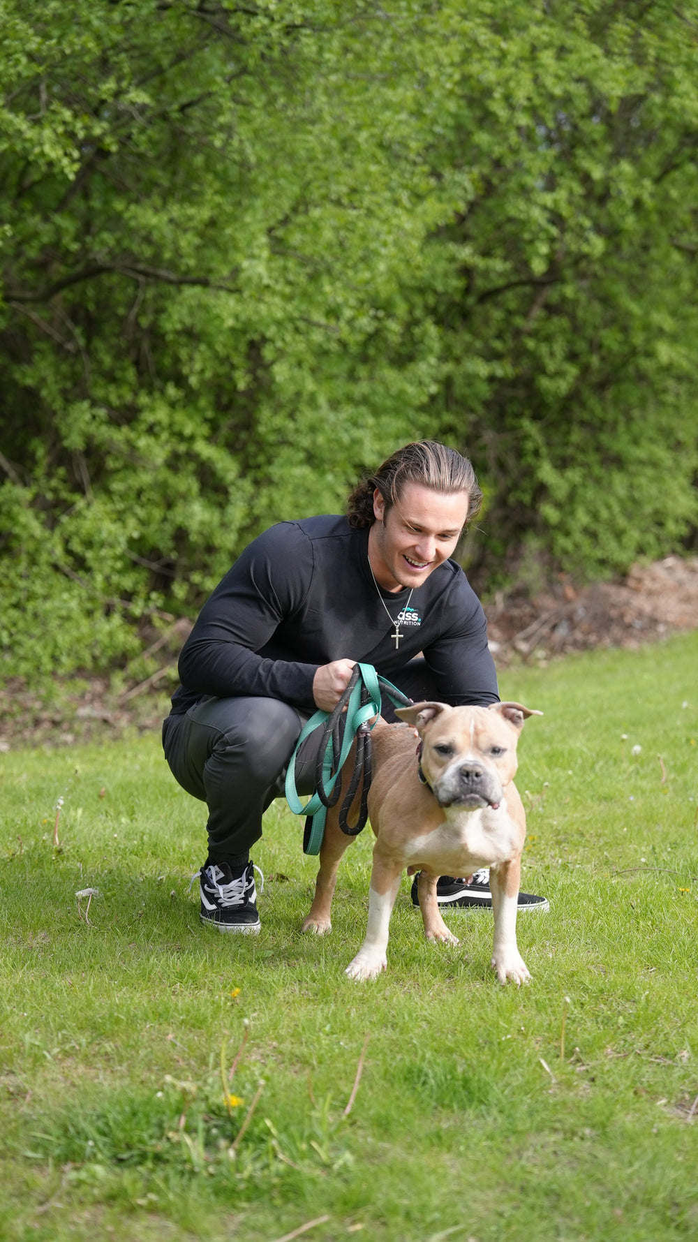 The founder kneeling on the grass beside a tan dog on a teal leash.