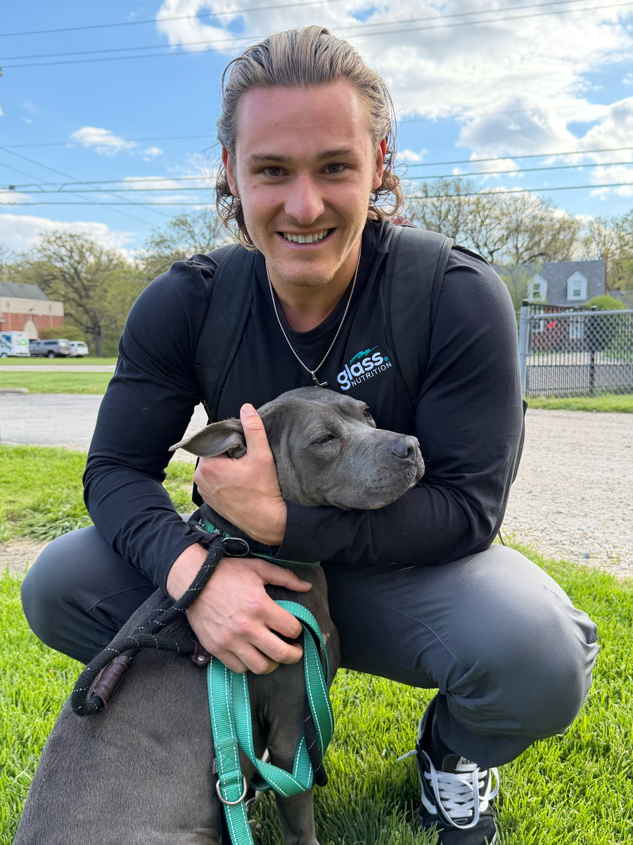 The founder kneeling on the grass with his arm around Raven, a gray pit bull on a teal leash.