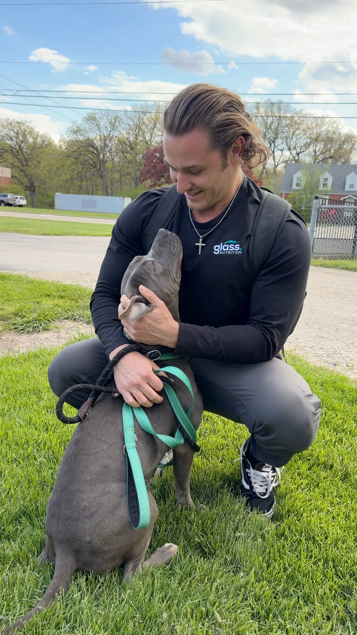 Gray pit bull on a teal leash nuzzling the face of a man kneeling on the grass.