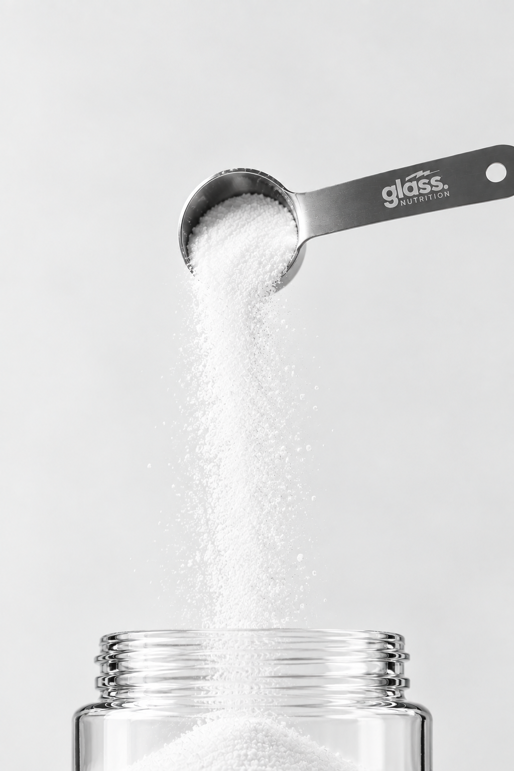A Glass Nutrition stainless steel scooper pouring creatine powder into an open glass jar